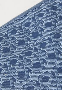 Tessuto blu con un intricato motivo tonale di lettere intrecciate, caratterizzato da una leggera lucentezza e una superficie liscia. Logo del marchio visibile.