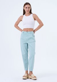 Lichtblauwe, hooglopende pantalon met een taps toelopende pijp, met voorzakken en een knoopsluiting. Gecombineerd met een witte mouwloze top.