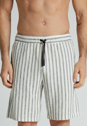 Męski tors w kremowych szortach z pionowymi szarymi paskami i czarnym sznurkiem, pokazujący dłonie i odkryty brzuch.