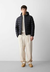 Giacca a vento nera con cappuccio, design a trapuntatura orizzontale, tasche frontali con zip. Indossata con un maglione crema e pantaloni chiari, scarpe beige.