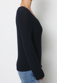 Kvinna klädd i en mörk marinblå ribbad stickad tröja med långa ärmar, tillsammans med ljusblå denimjeans, visad från sidan.