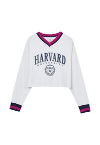 Felpa bianca corta con maniche lunghe, dotata di un colletto a V in rosa e blu navy. Sul fronte c'è la scritta "HARVARD UNIVERSITY" e l'emblema in blu navy.
