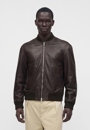 Homme portant une veste bomber en cuir brun foncé avec des poignets côtelés et un pantalon beige, debout devant un fond blanc uni.
