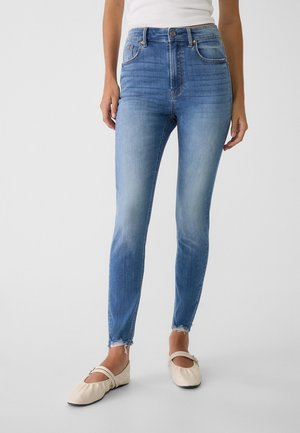 Kvinde iført lyseblå højtaljede skinny jeans med frynser i kanten og beige ballerinaer, stående mod en ensfarvet baggrund.