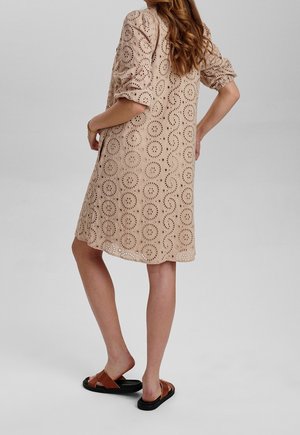 Vrouw met lang haar die een beige jurk met rond geperforeerd patroon en bruine sandalen draagt, staand met één hand op haar heup.