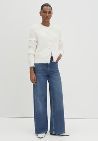 Cardigan blanc duveteux avec un col rond, une fermeture à boutons et des poignets côtelés, associé à un jean en denim bleu à jambe large et des bottines blanches.