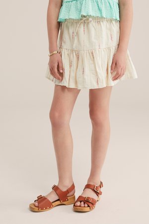 Underdel af en person iført en cremefarvet blomster nederdel, lysegrøn tekstureret top, et muslingearmbånd og brune læder sandaler med spænder og træsåler.