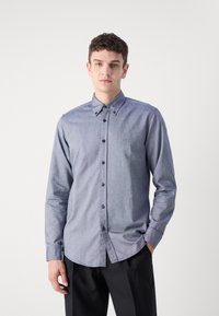 Light blue button-up shirt made of denim-like fabric, featuring a collar, front pocket detail, and dark buttons. Tapered fit with long sleeves.