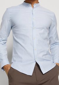 Camisa de botón azul claro con cuello alto, mangas largas y una textura de tela suave. Presenta costuras sutiles y un dobladillo redondeado.