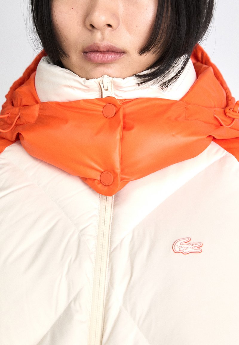 Giacca imbottita caratterizzata da un corpo in tessuto bianco e un cappuccio arancione, chiusura con zip e due bottoni decorativi sulla parte anteriore del cappuccio.