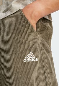 Corduroy pants in olive green with vertical ridges, featuring a front pocket and white embroidered Adidas logo on the lower leg.