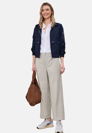 Kvinde iført navy jakke, hvid skjorte med knapper, beige bukser med vide ben, hvide sneakers og holder en brun ruskindstaske, smilende.