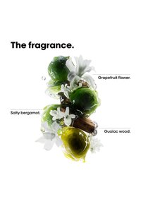 Frutti verdi di bergamotto, fiori bianchi di pompelmo e pezzi di legno di guaiaco circondati da gocce di liquido trasparente su sfondo bianco.