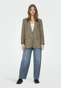 Blazer verde oliva oversize con un bottone e due tasche frontali, abbinato a jeans a gamba larga blu chiaro e scarpe con zeppa nere.
