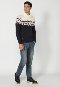 Jersey de punto azul marino con un cuello en crema y rojo, que presenta un diseño en la parte superior. Llevado con vaqueros azules y zapatillas negras.