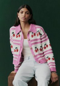 Gebreid vest in roze en wit, met rode kersenpatronen, contrasterende strepen en houten knopen. Zachte textuur met een kraag.