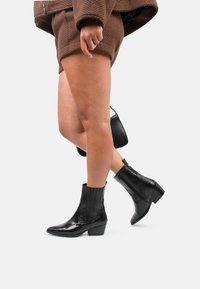 Botas negras hasta el tobillo con puntera puntiaguda, superficies texturizadas, un tacón bajo en bloque y estrías horizontales en el caña. Acabado ligeramente brillante.