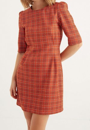 Kvinde iført en tætsiddende, knælange orange kjole med hvidt ternmønster og albu-lange ærmer, stående foran en ensfarvet baggrund.