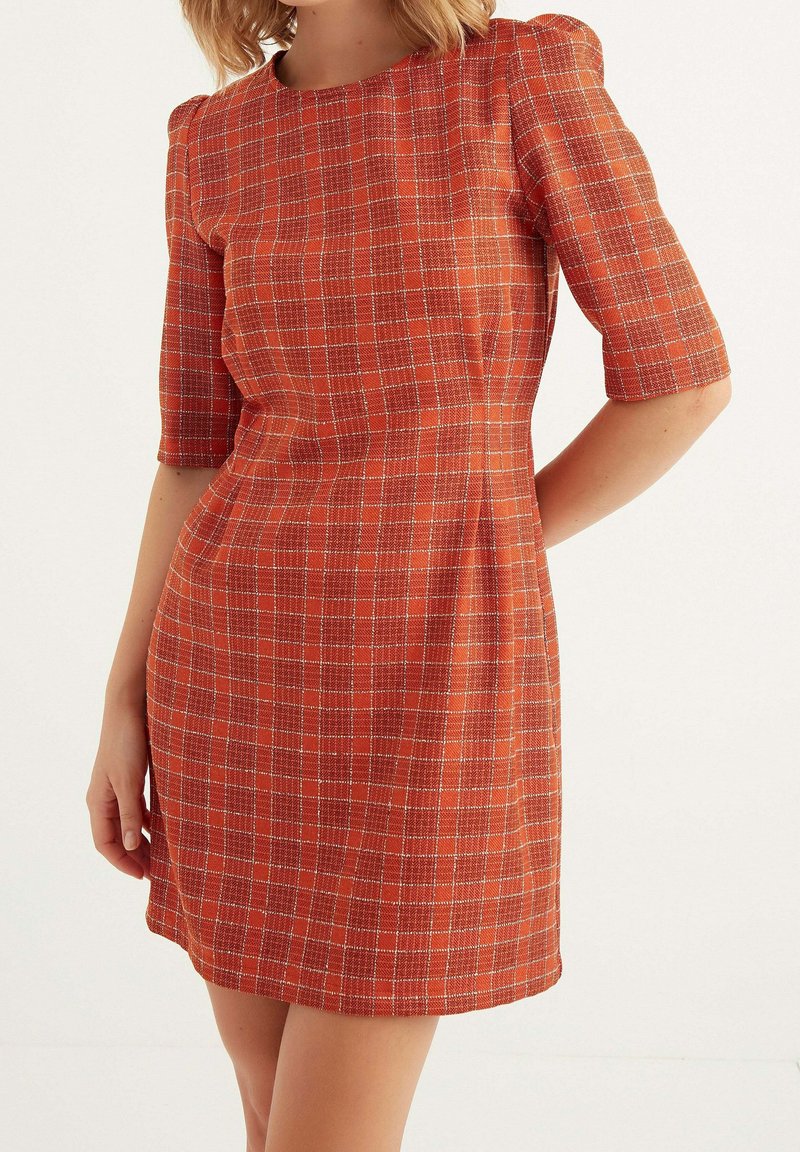 Femme portant une robe orange ajustée, longueur genou, avec un motif à carreaux blanc et des manches jusqu'aux coudes, debout devant un fond uni.