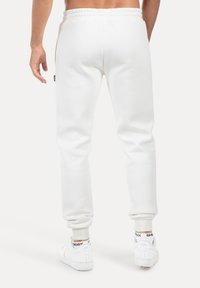 Pantalones joggers de forro polar blanco con cintura y puños elásticos, con bolsillos laterales y un pequeño parche de logo negro. Llevados con zapatillas blancas.