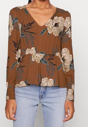 Top lungo a maniche con stampa floreale marrone, scollatura a V, con vita a peplo e grandi fiori color crema accentuati da foglie verdi. Abbinato a jeans chiari.