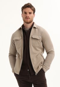 Hombre con chaqueta beis de cremallera encima de una camisa y pantalones marrón oscuro, de pie con las manos en los bolsillos frente a un fondo blanco.