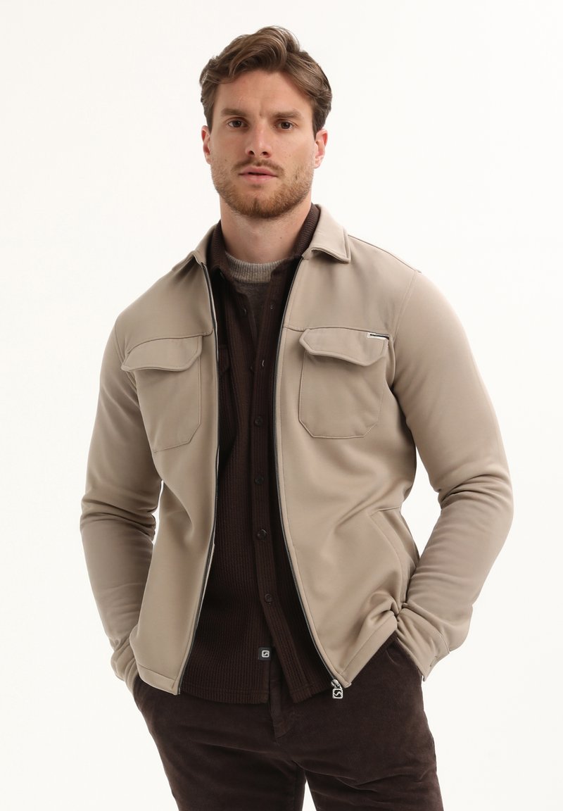 Hombre con chaqueta beis de cremallera encima de una camisa y pantalones marrón oscuro, de pie con las manos en los bolsillos frente a un fondo blanco.