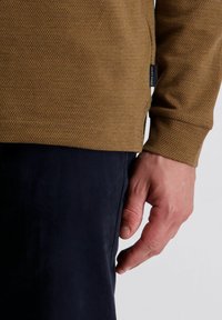 Brauner Pullover mit strukturiertem Muster, mit langen Ärmeln und einem Seitenetikett, kombiniert mit dunklen marineblauen Hosen. Die Hand ruht an der Seite.