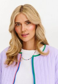 Blondynka z falującymi włosami nosi pastelowy fioletowy żakiet z zielonym i niebieskim wykończeniem na białej koszuli oraz kolorową naszyjną z koralików.