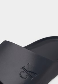 Μαύρο σανδάλι slide Calvin Klein με ανάγλυφο λογότυπο "cK" σε φαρδιά λουράκι, απεικονιζόμενο σε κοντινή λήψη σε λευκό φόντο.