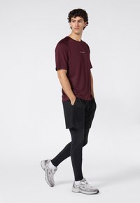 Młody mężczyzna ubrany w bordową koszulę, czarne szorty, czarne legginsy i białe sneakersy, stojący z rękami w kieszeniach na białym tle.