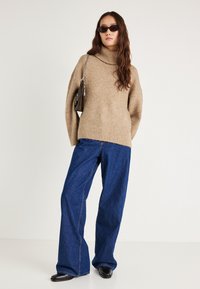 Šķiedraina bēša augstās apkakles džemperis ar ribotu apakšmali, valkāts kopā ar ziliem plašām kāju biksēm. Modelis rokās tur melnu rokassomu ar ķēdes siksnu, uzsaucot saulesbrilles.