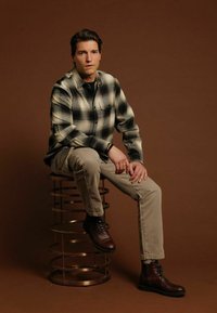 Zwart-wit geblokte flanellen shirt, beige broek, bruine lederen laarzen, zittend op een gouden metalen kruk tegen een bruine achtergrond.