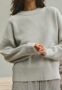 Pull gris clair côtelé avec une coupe décontractée, des poignets larges et un col rond. Le tissu texturé contraste avec une bande horizontale lisse à la taille.
