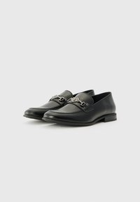 Mocasines de cuero negro con un diseño elegante, que presentan un detalle decorativo en forma de hebilla de plata, costuras decorativas y un tacón bajo.