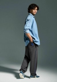 Camicia di jeans azzurro chiaro, pantaloni tuta neri con accenti blu e sneakers navy. Modello di profilo, mani in tasca. Sfondo semplice.