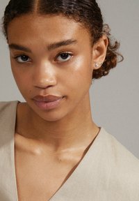 Gros plan sur un visage avec une peau brun moyen, portant des vêtements beige clair. Les caractéristiques notables incluent de petites boucles d'oreilles en argent en forme de cœur.