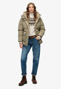 Chaqueta acolchada verde oliva con capucha, suéter beige con estampado y jeans azules. Botas marrones con cordones completan el conjunto.