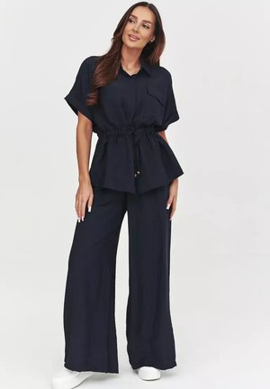 Kobieta stojąca z rękami w kieszeniach, ubrana w granatowy krótki rękaw z paskiem i szerokie spodnie z białymi trampkami na białym tle.