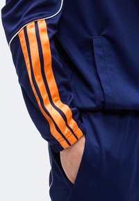 Άτομο με ναυτικό μπλε φόρμα με τρεις πορτοκαλί ρίγες στο μανίκι, με το χέρι άνετα στην τσέπη του παντελονιού.