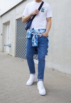 Biała koszulka z logo Levis, niebieski sweter tie-dye związany w talii, niebieskie dżinsy i białe sneakersy na teksturowanym chodniku.