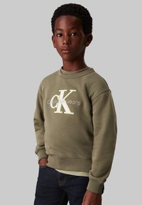 Felpa in cotone verde oliva con collo rotondo, caratterizzata da un grande logo ricamato bianco e giallo sul davanti. Indossata con jeans blu scuro.