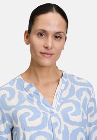 Camicia blu e bianca con un motivo vorticoso, caratterizzata da una scollatura a V e una consistenza del tessuto morbida. Il design è rilassato e informale.