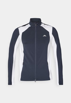 Marineblå og hvit atletisk jakke med høy krage, hel glidelås, lange ermer og glidelåslommer i sidene; har logo på brystet.