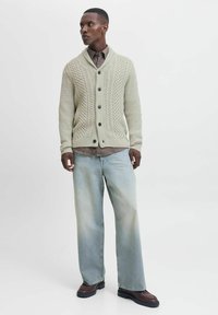 Cardigan en tricot vert clair avec un motif en câble, six boutons et un col châle, associé à un jean en denim bleu clair ample et des chaussures marron.