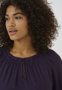 Purple blouse with a gathered round neckline, small keyhole detail, and subtle pleating. The fabric appears smooth with a soft texture.