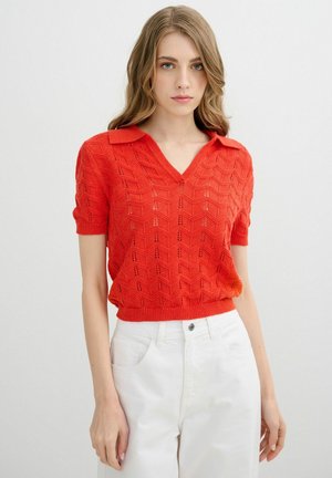 Mujer con un top rojo de punto de manga corta y cuello, combinado con pantalones blancos de talle alto, posando frente a un fondo liso.
