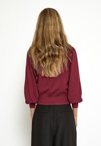 Burgunderfarbener Pullover mit Ballonärmeln und geripptem Saum, kombiniert mit schwarzen, hoch taillierten Hosen. Glatte Textur und figurbetontes Design hervorgehoben.
