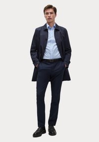 Marineblauer Trenchcoat mit Knöpfen, getragen über einem hellblauen Hemd und dunklen Hosen, kombiniert mit schwarzen Schuhen, zeichnet sich durch ein maßgeschneidertes Design aus.