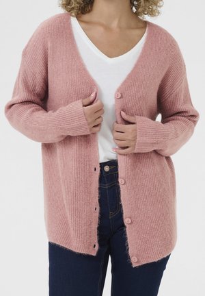 Personne portant un cardigan rose doux boutonné sur un t-shirt blanc à col en V et un jean bleu foncé ajustant les manches du cardigan.